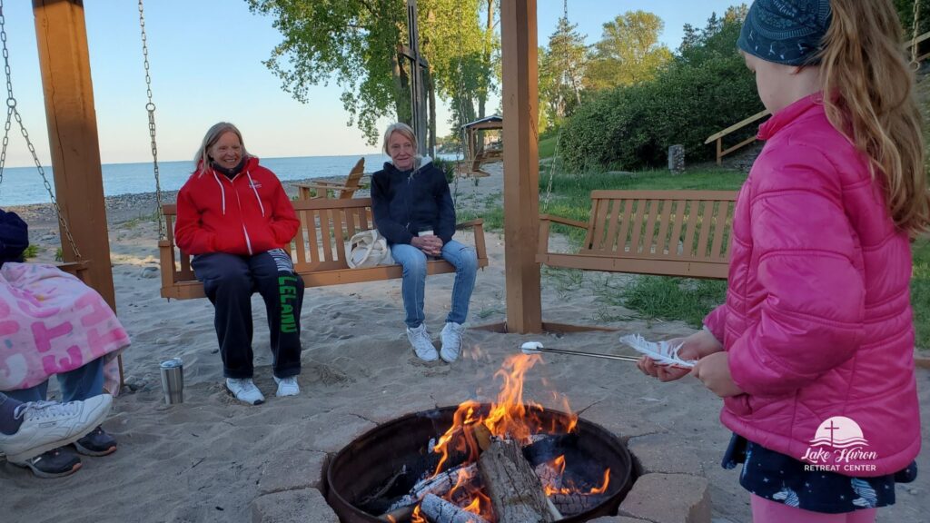 Adults on beachside swings while child roasts a marshmallow over a campfire.