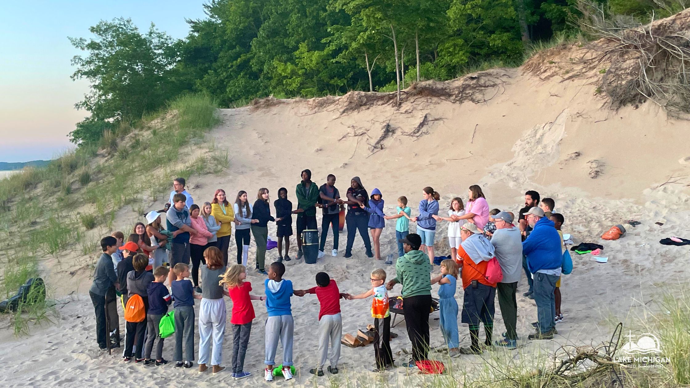 Children of all ages and races join hands with each other and adult counselors on the sand near the shore of Lake Michigan and Lake Michigan Camp & Retreat.
